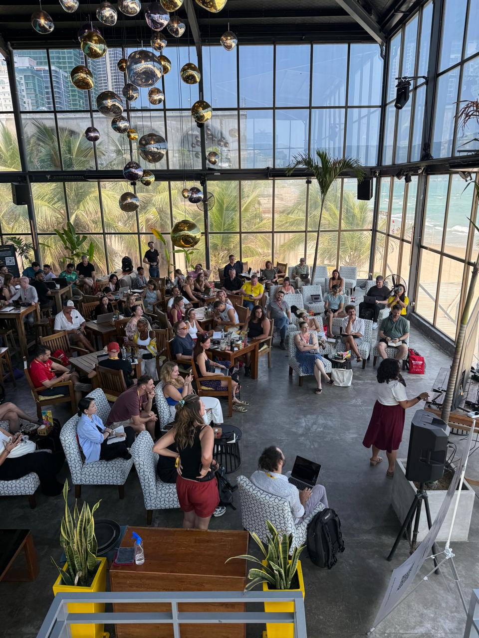 Full house at ESCO Beach Da Nang — sea-view rooftop, panorama windows, palms outside, presenter on stage.